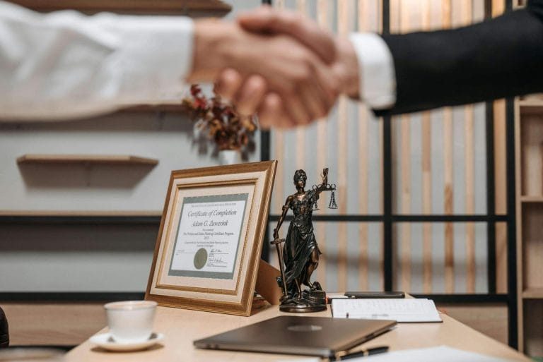 Legal office desk with Lady Justice statue, framed certificate, and people shaking hands in the background.