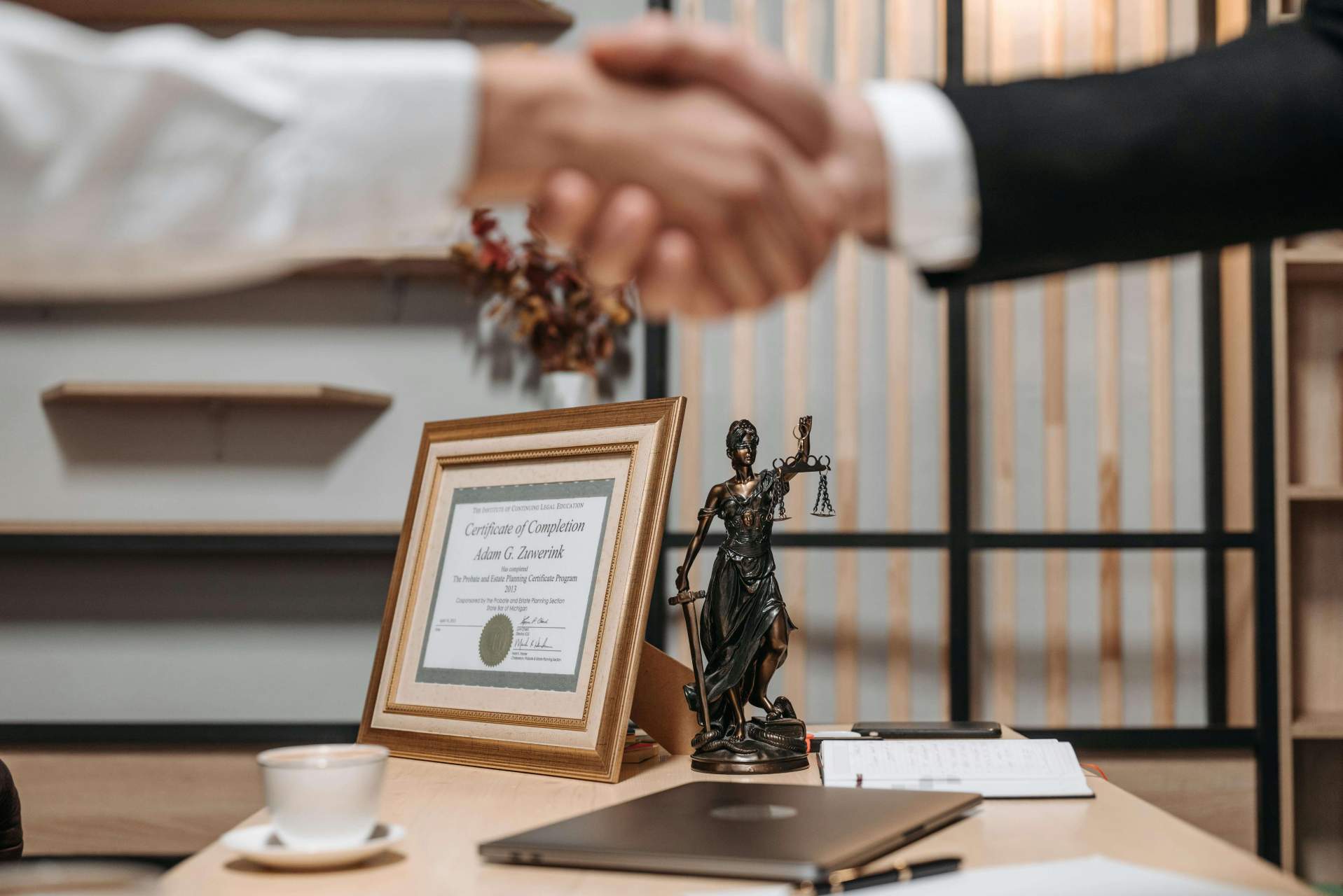 Legal office desk with Lady Justice statue, framed certificate, and people shaking hands in the background