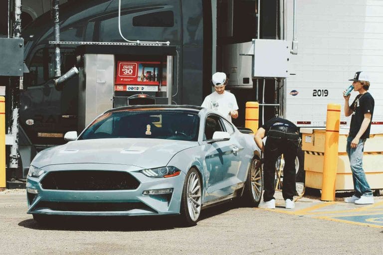 A silver sports car parked at a gas station while three people stand nearby, including one person fueling the car, with a large truck and fuel pumps in the background