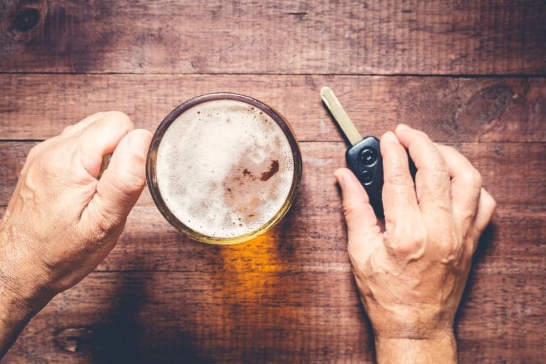 Person holding a beer glass near car keys on a wooden table, suggesting the risk of drinking and driving.
